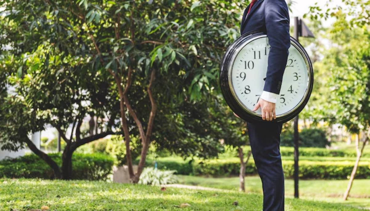 A businessman in a suit stands in a park, holding a large wall clock, symbolizing time management.
