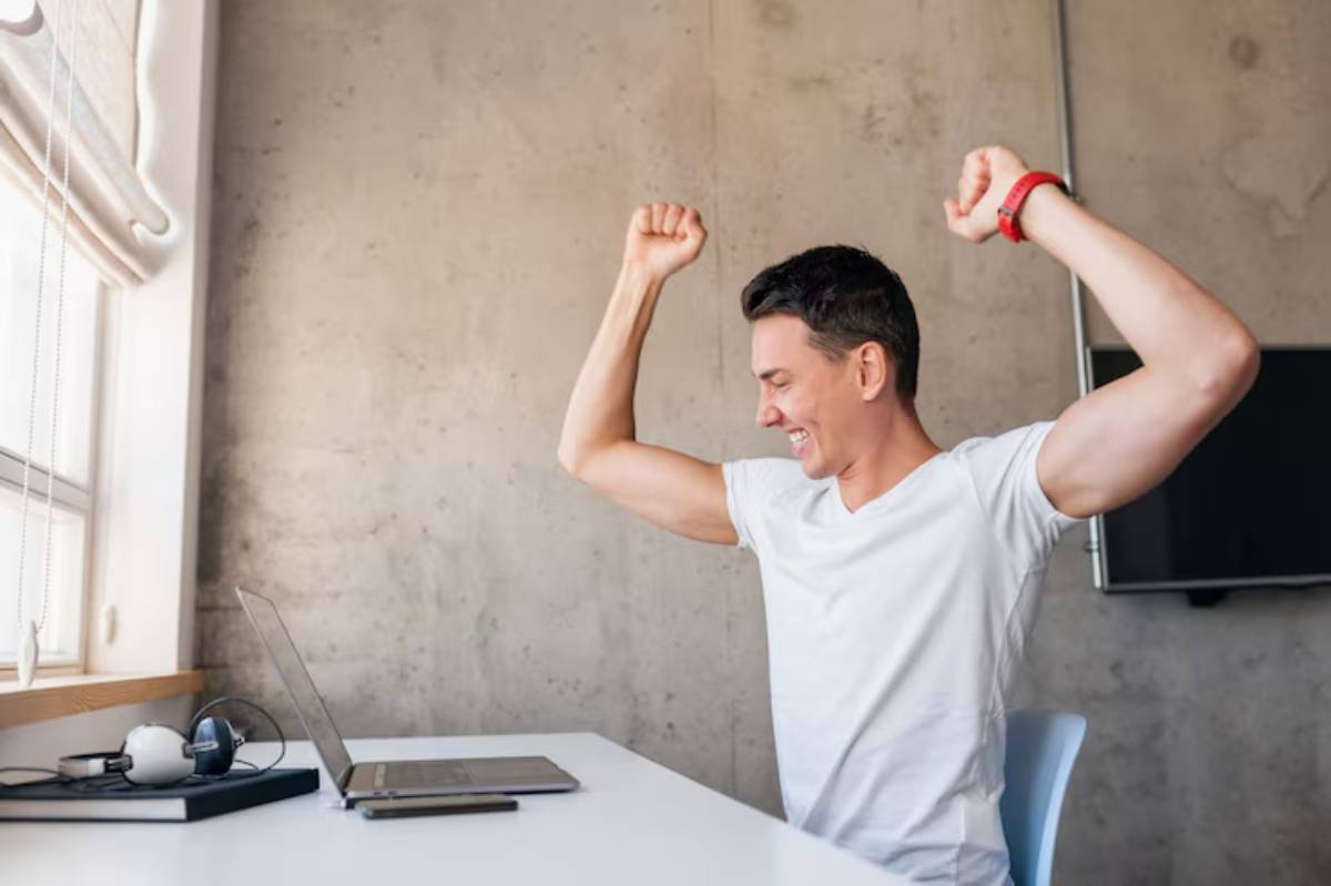 A person in a white t-shirt celebrates success at a desk with a laptop, arms raised in triumph, in a modern workspace.