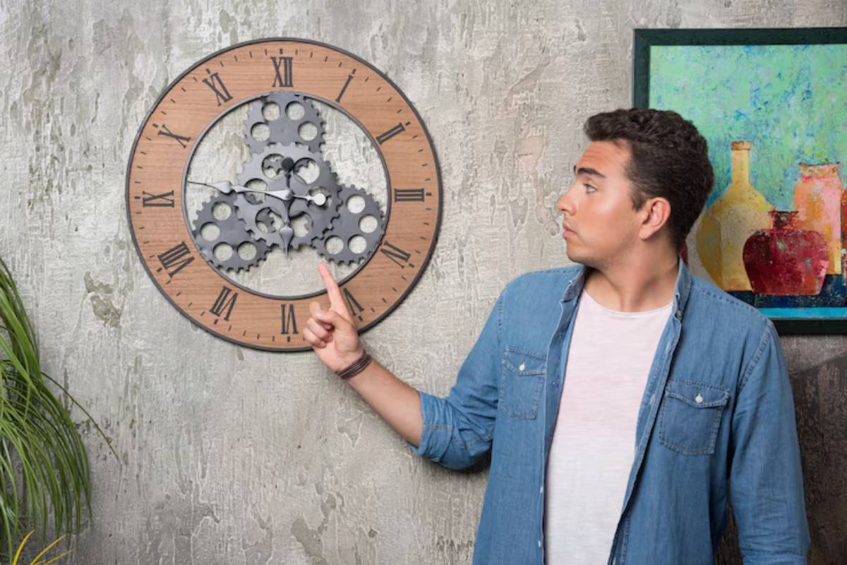 A young man in a denim shirt points at a unique wall clock with visible gears and Roman numerals, set against a textured wall.