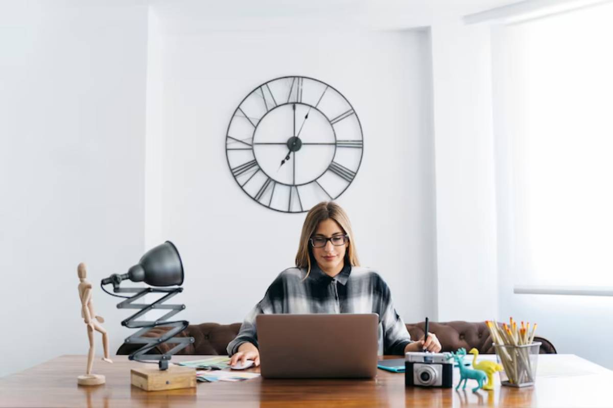 A woman works at a desk with a laptop, surrounded by stationery, a lamp, and decorative figures, with a large clock on the wall behind her.