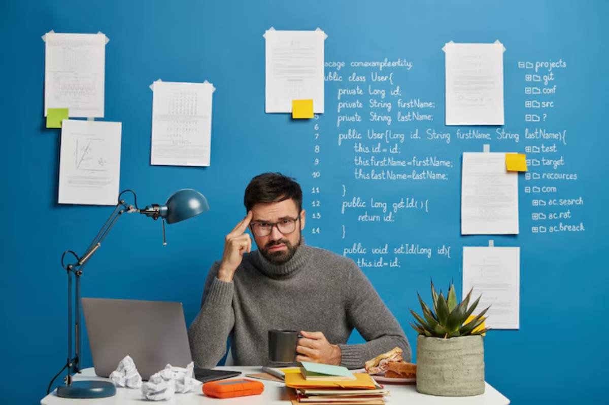 A person in a gray sweater sits at a desk with a laptop, papers, and a plant against a blue wall covered in notes and code snippets.