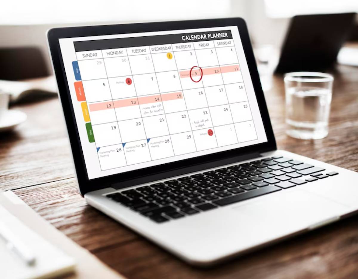 A laptop displays a colorful calendar planner on a wooden table, with a cup of water nearby, emphasizing productivity and organization.