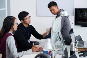 Three people engage in discussion around an office desk with computers, coffee cups, and various office supplies.