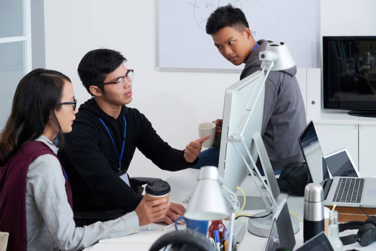 Three people engage in discussion around an office desk with computers, coffee cups, and various office supplies.