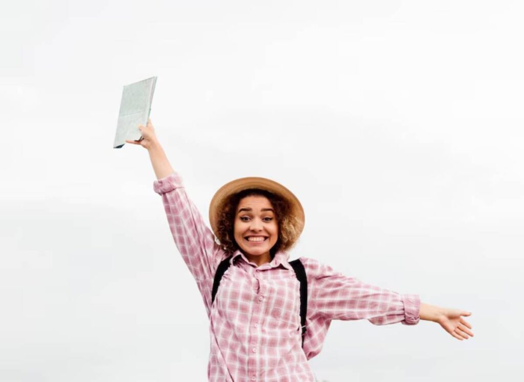 A person in a pink plaid shirt and straw hat joyfully holds a book aloft against a bright, cloudy sky.