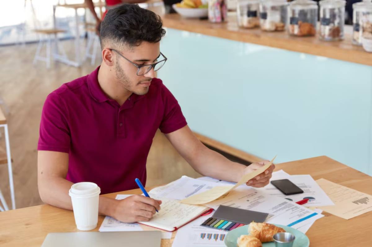 A person in a red polo shirt sits at a wooden table covered with papers, a notebook, a phone, and pastries, focused on their notes.