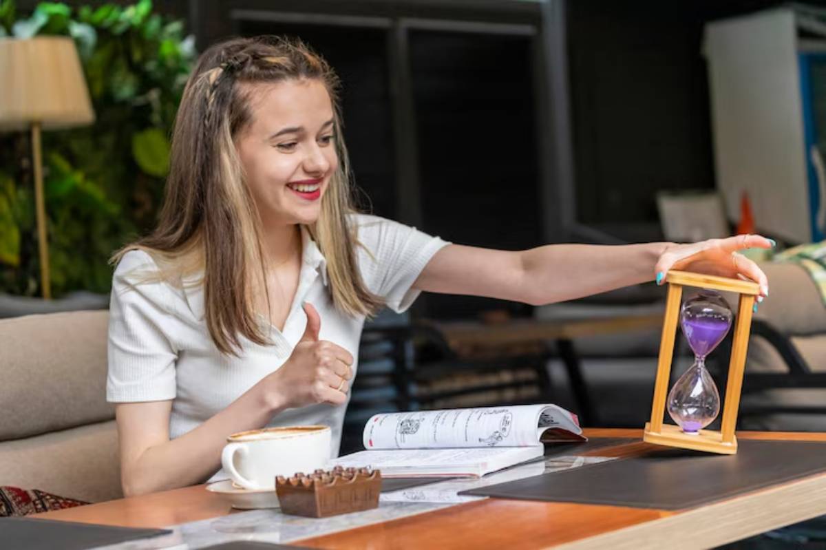 A person sits at a table with a cup of coffee, holding an hourglass with purple sand, thumbs-up beside an open book.