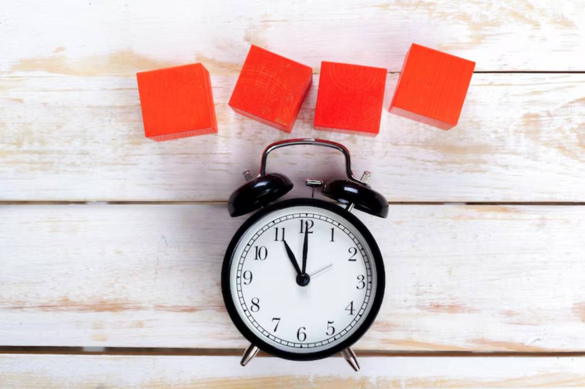 A black alarm clock showing 10:10, surrounded by four bright red wooden blocks on a white wooden surface.