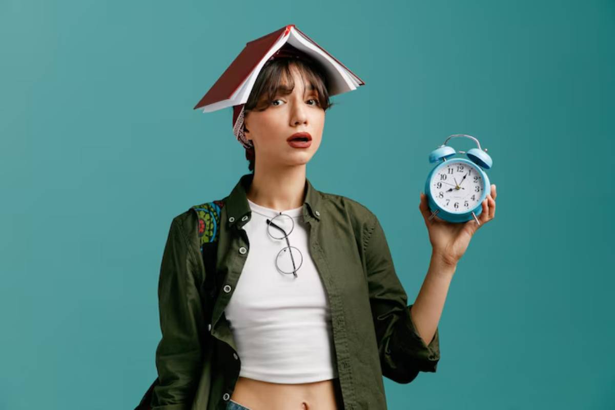 A person wears a book as a hat and holds a vintage blue alarm clock against a teal background.