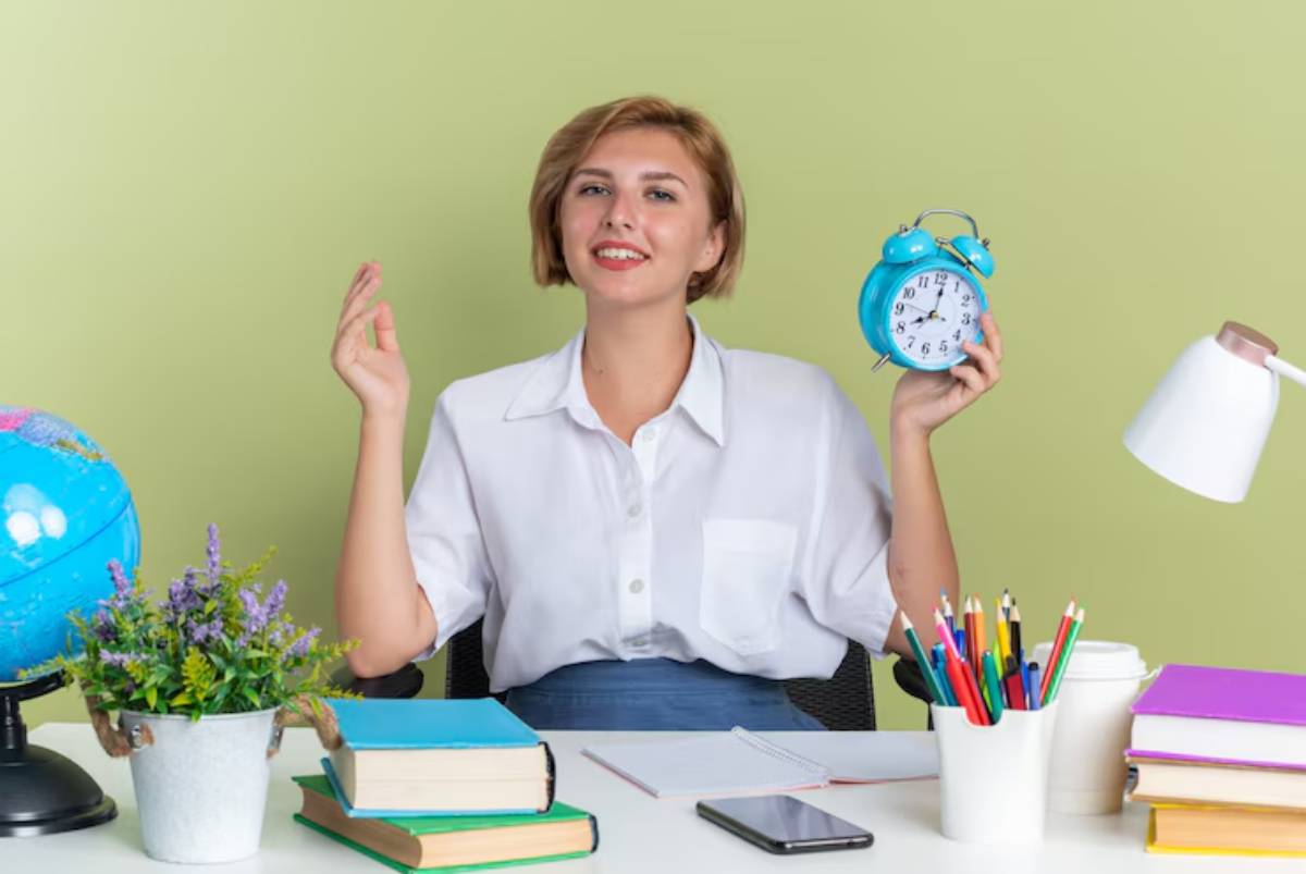 A person holds a blue alarm clock, surrounded by books, a globe, and colorful pens, sitting at a desk against a green background.