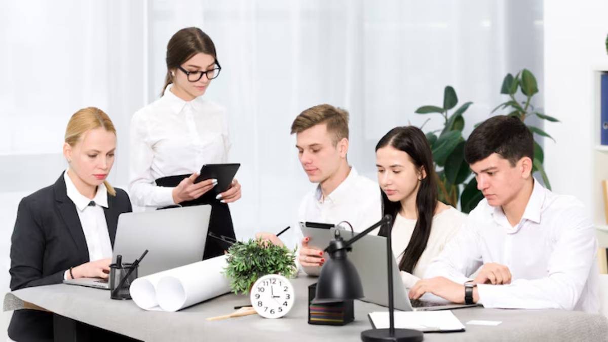 A group of professionals collaborate around a table with a laptop, tablets, and a clock, in a modern office setting.