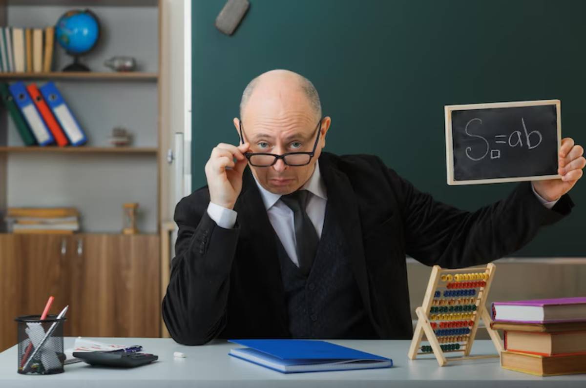 A man in formal attire sits at a desk, holding a small chalkboard with an equation, surrounded by books and educational tools.