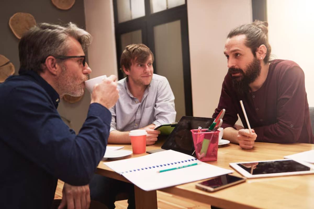 A group of men engage in a brainstorming session around a wooden table, with coffee cups and tablets present. Notes and a pen are visible.